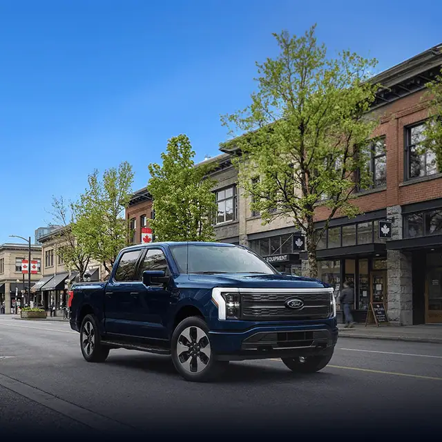 Blue Ford pickup truck driving through a quaint town street lined with shops and trees.
