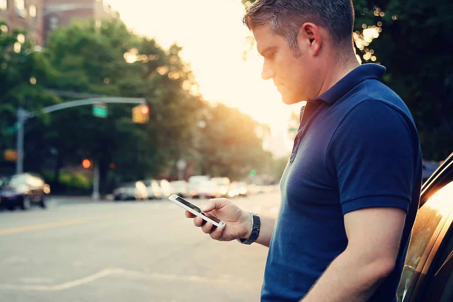 Homme en chemise bleue utilisant un smartphone dans une rue de la ville au coucher du soleil. Concept de mode de vie urbain.